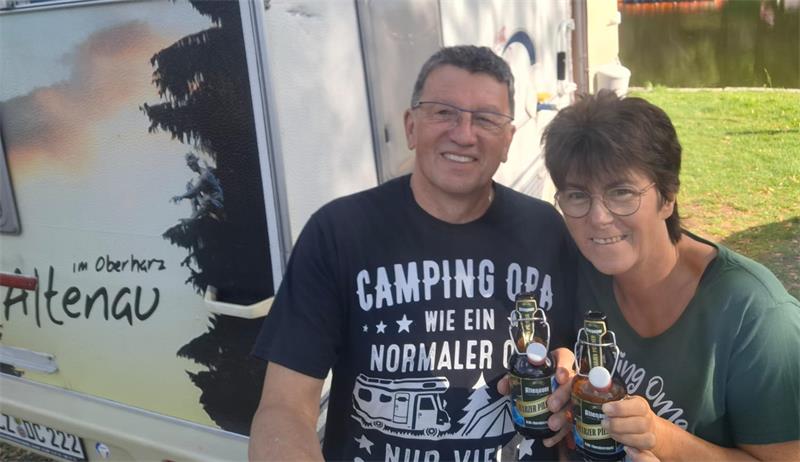 Mann und Frau stehen lächelnd vor einem Wohnwagen und halten jeweils eine Flasche in der Hand.