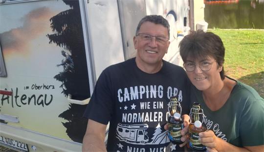Mann und Frau stehen lächelnd vor einem Wohnwagen und halten jeweils eine Flasche in der Hand.