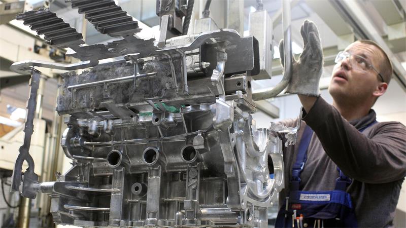 Mann mit Schutzbrille und Handschuhen inspiziert großen Metallmotorblock in Werkhalle.