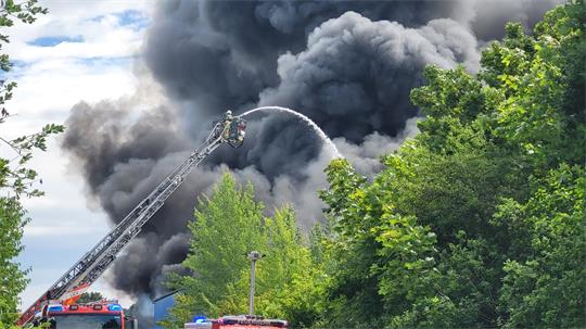 Das Bild zeigt einen Feuerwehrmann auf der Drehleiter beim Löschen der Lagerhalle mitten in einer mächtigen Raucwolke.