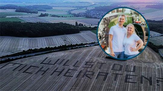 Landschaft mit Feldern und einem großen Schriftzug in den Feldern, der 'WILLST DU MICH HEIRATEN' bildet.