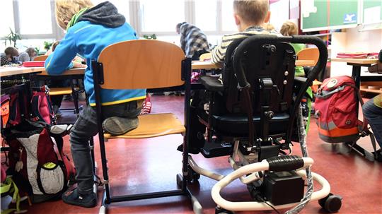 Ein Kind mit Behinderung sitzt in seinem Rollstuhl gemeinsam mit anderen Erstklässlern ohne Handicap in einem Klassenraum. Inklusion soll Teilhabe für Menschen mit Beeinträchtigung ermöglichen.