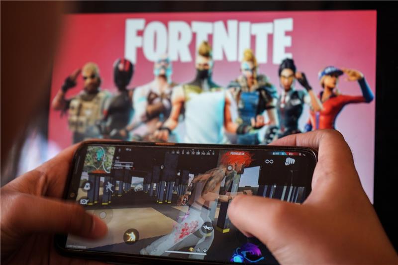 Videospiele: Wann muss ich mir Sorgen machen? Ein Kind spielt ein Spiel auf einem Mobiltelefon mit einem Bild des Fortnite-Spiels auf dem Computerbildschirm im Hintergrund. Foto: picture alliance/dpa/ZUMA Wire | Herwin Bahar