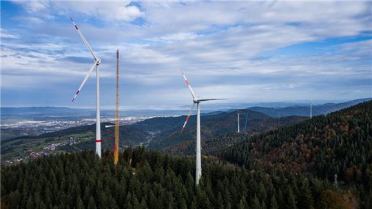 Ein Kran zieht das dritte Rotorblatt einer neuen Windkraftanlage auf der Holzschlägermatte am Schauinsland hoch während daneben eine kleinere Windkraftanlage und im Hintergrund eine weitere im Bau befindliche Windkraftanlage zu sehen sind. Windkraft im Wald ist höchst umstritten.