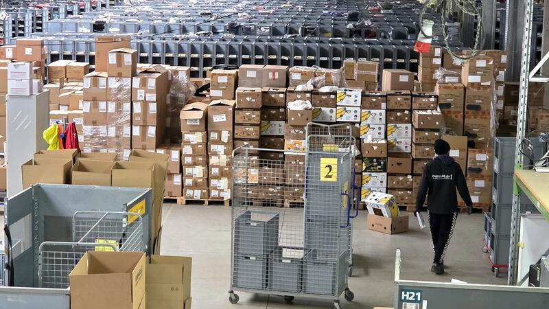 Lagerhalle mit hohen Regalen, vielen gestapelten Kartons und einem Mitarbeiter, der an den Kartons vorbeigeht