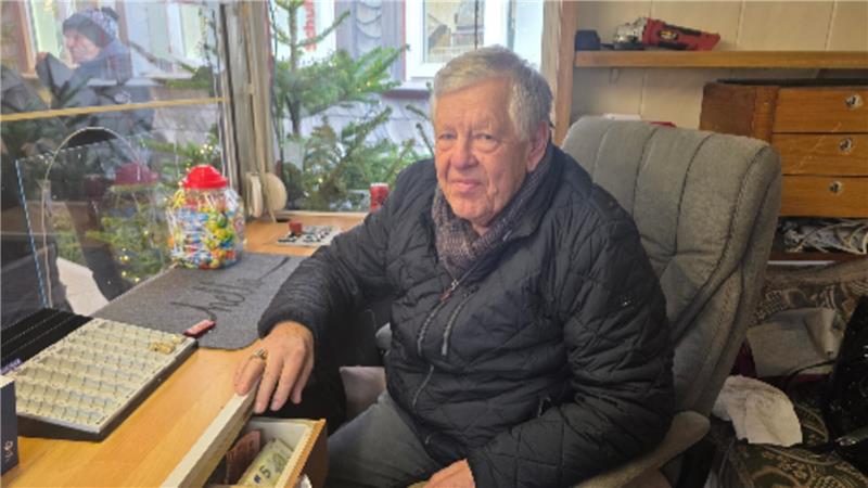 Mann mit grauem Haar und schwarzer Steppjacke sitzt an einem Schreibtisch mit geöffnetem Schublade und einem Spielbrett mit weißen und schwarzen Steinen.