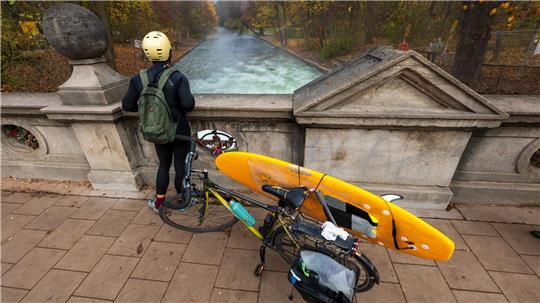 Ein Mann mit einem Surfboard an seinem Fahrrad schaut von einer Brücke auf die - nicht mehr vorhandene - Eisbachwelle im Englischen Garten.