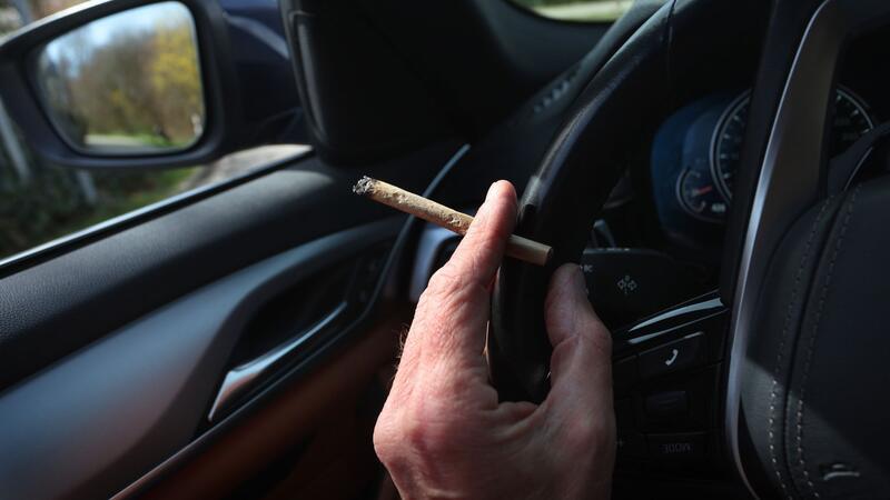Innenministerin gegen höheren Cannabis-Grenzwert im Verkehr Ein Mann sitzt mit einem Joint zwischen den Fingern am Steuer eines Autos.