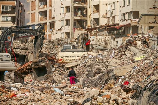 Ein Mann steht in Samandag in der Türkei zwischen Trümmern, während Bagger die Überreste eines zerstörten Gebäudes abtransportieren. Nach dem katastrophalen Beben am 6.2. kommt es in der Region immer wieder zu Nachbeben.