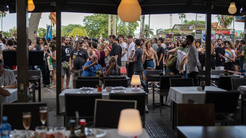 Ein Mann und eine Frau auf der Terrasse eines Restaurants in Barcelona lassen sich von vorbeiziehenden Demonstranten gegen den Massentourismus offenbar nicht aus der Ruhe bringen.