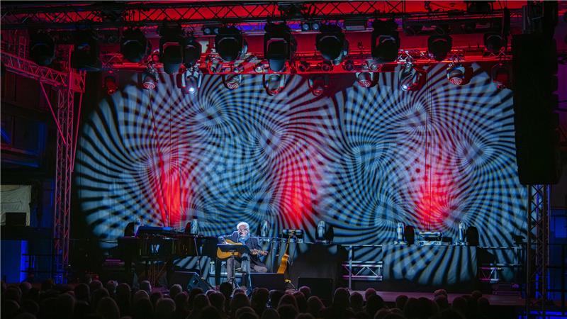 Heinz Rudolf Kunze präsentiert in Goslar ein intensives Solo Die rot-blau beleuchtete Bühne mit Heinz Rudolf Kunze, der mit Gitarre in der Mitte sitzt und singt.