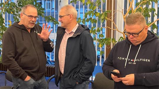 Drei Männer in Freizeitkleidung stehen in einem Raum mit großen Fenstern und Pflanzen, zwei unterhalten sich, einer schaut auf sein Smartphone