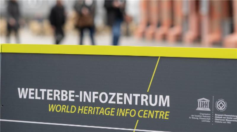Schild mit der Aufschrift 'WELTERBE-INFOZENTRUM WORLD HERITAGE INFO CENTRE' vor unscharfem Hintergrund mit Personen und Gebäuden