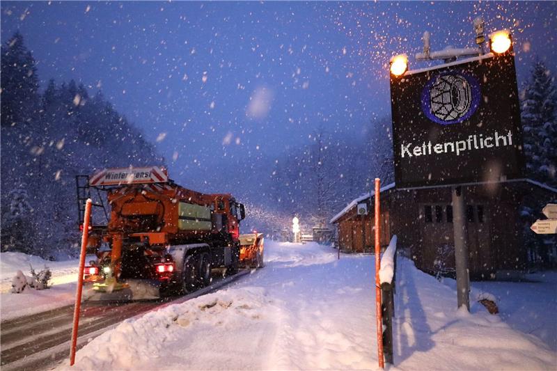 Schneefall in Bayern - erhebliche Lawinengefahr in den Alpen Ein Schild weist auf die Schneekettenpflicht am Riedbergpass im bayerischen Landkreis Oberallgäu hin, während daneben ein Räumfahrzeug vom Winterdienst den Schnee von der Straße räumt. (Symbolbild)