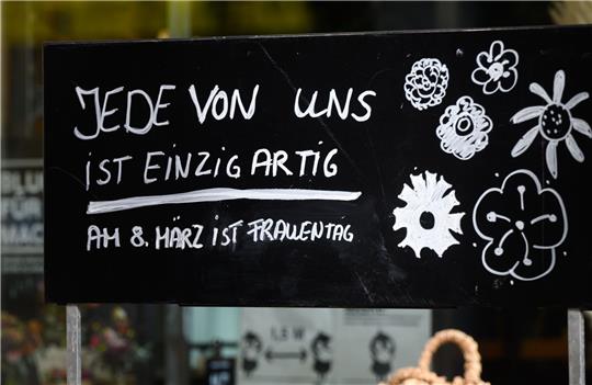 Ein Schriftzug „Jede von ist einzigartig - Am 8. März ist Frauentag“ steht auf einem Schild vor einem Blumenladen.