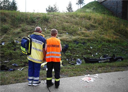 Ein Seelsorger und ein Mitglied der Feuerwehr an einer Unfallstelle auf der Autobahn 9 vor einer Böschung, die mit Trümmerteilen übersäht ist.