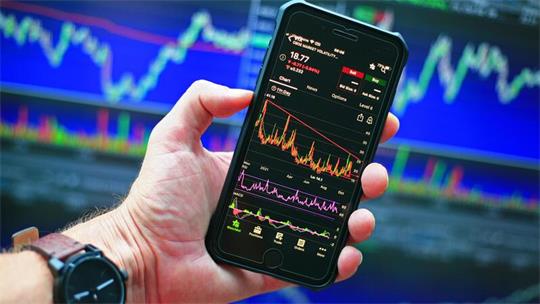 Hand hält Smartphone mit Finanzdiagrammen und Kursdaten vor unscharfem Bildschirm mit Börsengrafiken