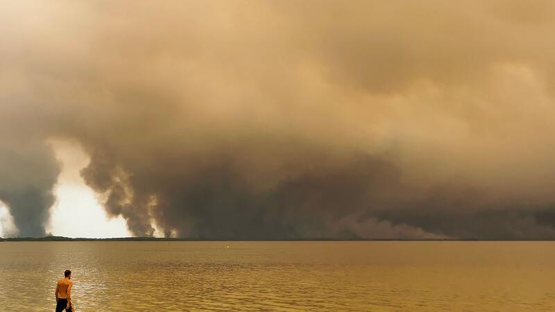 Ein Mann am Wasser, dahinter steigen Rauchwolken über dem Meer auf.
