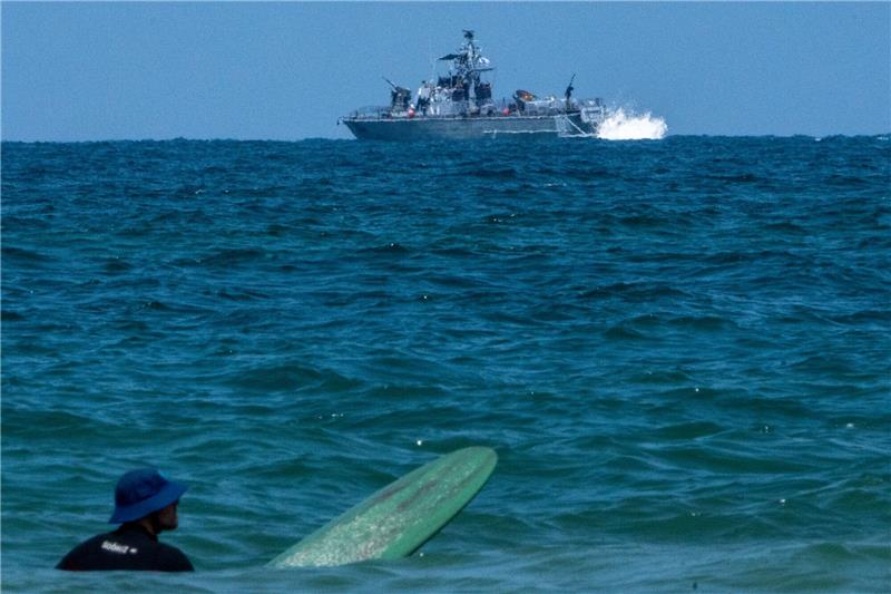 Ein Surfer wartet auf eine Welle, während ein israelisches Marineschiff im Mittelmeer vor der Küste von Hadera patrouilliert.