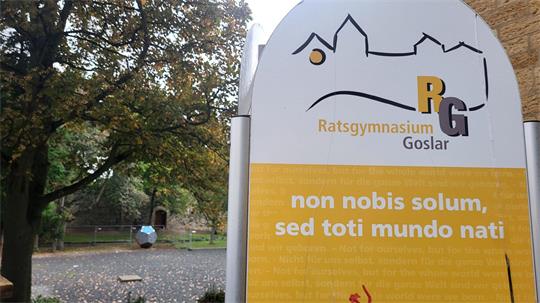 Schild mit der Aufschrift 'Ratsgymnasium Goslar' und lateinischem Text vor einem von Bäumen umgebenen Schulgelände