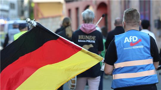 Ein Teilnehmer der Montagsdemonstration in Halberstadt trägt eine gelbe Warnweste mit einem AfD-Logo und der Aufschrift "Deutschland. Aber normal." und hält eine Deutschlandfahne. Seit Dezember 2021 finden sich die Teilnehmer regelmäßig zusammen.