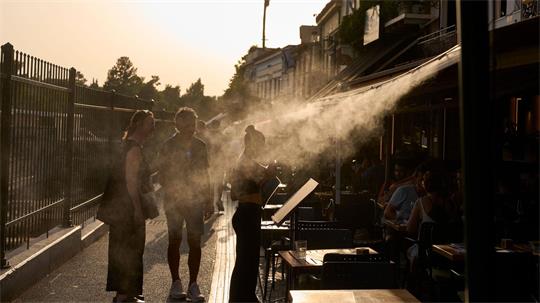 Ein Touristenpaar geht an einer Sprühanlage vorbei, während ein Restaurantmitarbeiter sie im Athener Stadtteil Monastiraki zu ihrem Tisch führt.