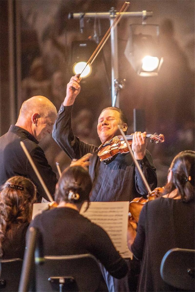 Ein Konzet im Gegenlicht zweier Scheinwerfer: In der Mitte einiger Musiker ein Geiger, der den Geigenbogen in die Luft schwingt und lacht. 