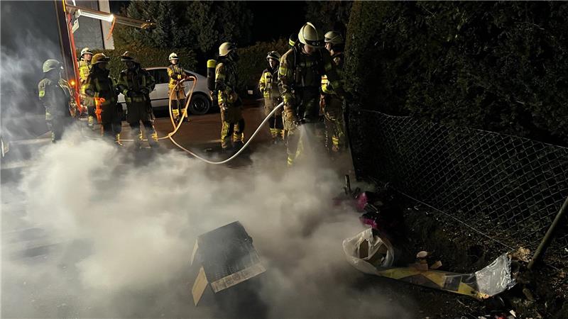 Das Foto zeigt Einsatzkräfte der Feuerwehr, die in der Robert-Koch-Straße einen brennenden Zaun löscht.