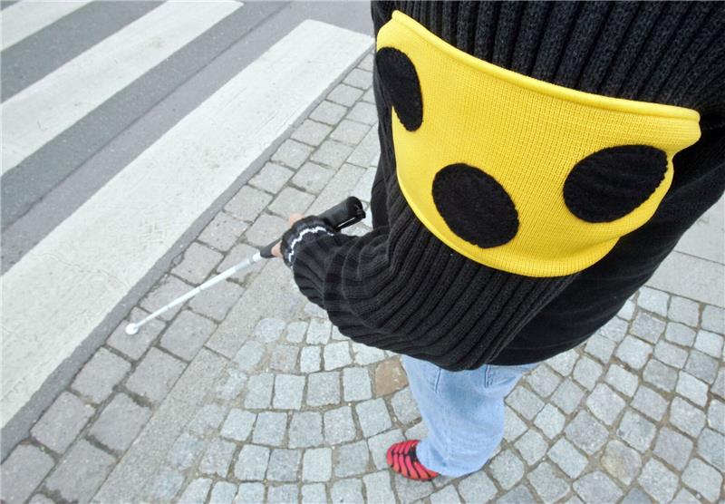 Sehbehindertenverband: Besser wahrnehmbare E-Auto-Geräusche Ein blinder Mann geht mit seinem weißen Taststock und einer Armbinde mit drei Punkten über eine Straße in Hannover.