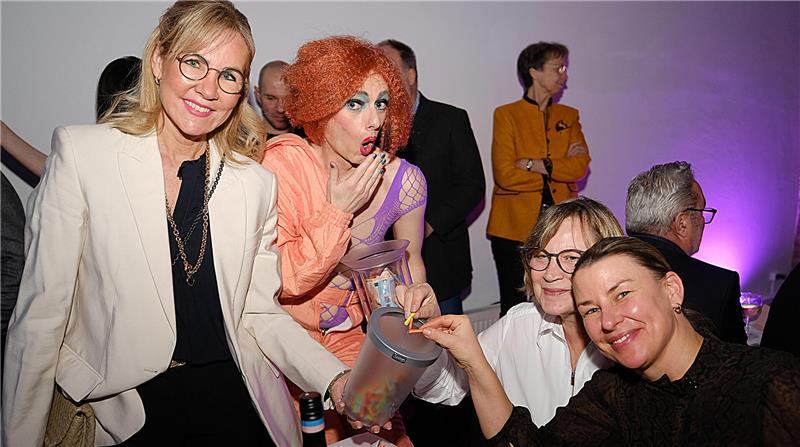 Eine Frau mit Losbehälter lächelt in die Kamera, neben ihr macht eine Drag-Künstlerin Dönekens, zwei Frauen ziehen Lose. 