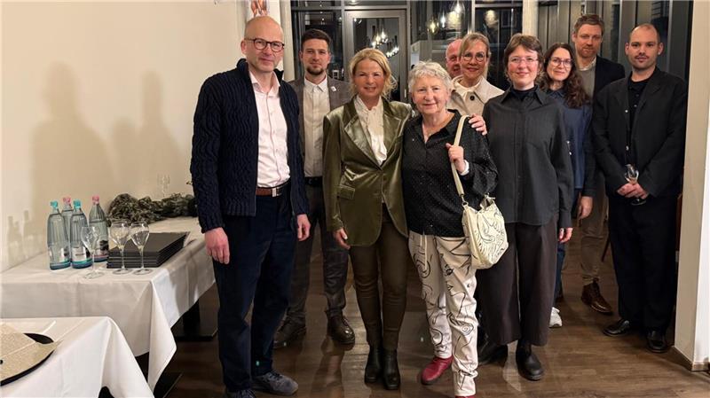Gruppe von zehn Personen in einem Raum mit Holzboden und weißen Wänden, links ein Tisch mit Wasserflaschen und Gläsern.