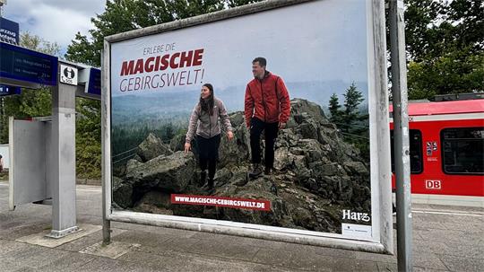 Das Foto zeigt an einer S-Bahn-Haltestelle ein großflächiges Werbeplakat mit einer Frau und einem Mann auf einem Felsen. Auf dem Plakat steht "Erlebe die magische Gebirgswelt!"