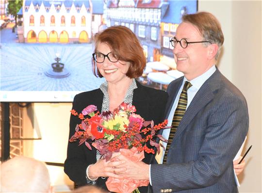 Zwei Personen in formeller Kleidung, eine hält einen Blumenstrauß, im Hintergrund ein Bildschirm mit einem Stadtbild.