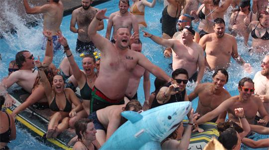 Gruppe von Menschen in Badebekleidung, die in einem Pool jubeln und eine aufblasbare Haifigur halten.