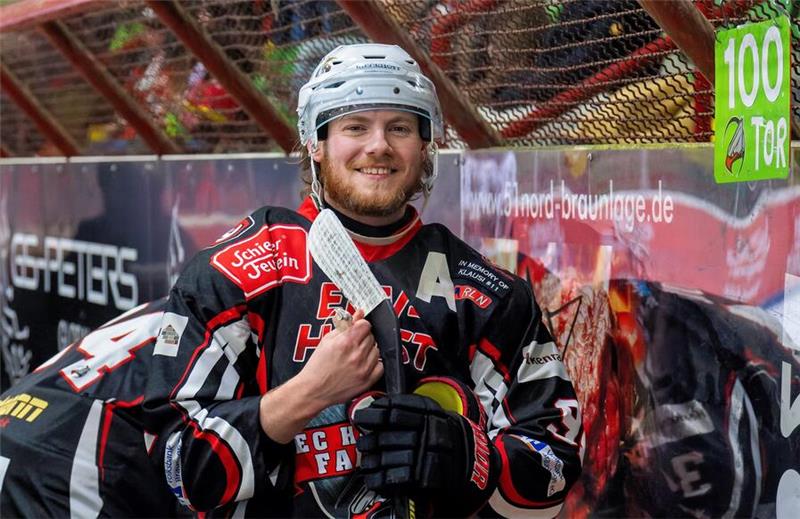 Der Eishockeyspieler der Harzer Falken, Regan Nagy steht in voller Montur und Schläger in der Hand vor der Tribüne.