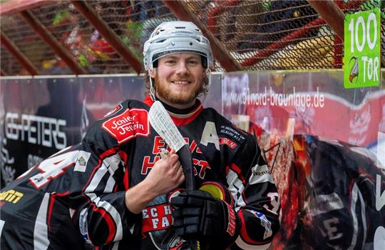 Der Eishockeyspieler der Harzer Falken, Regan Nagy steht in voller Montur und Schläger in der Hand vor der Tribüne.