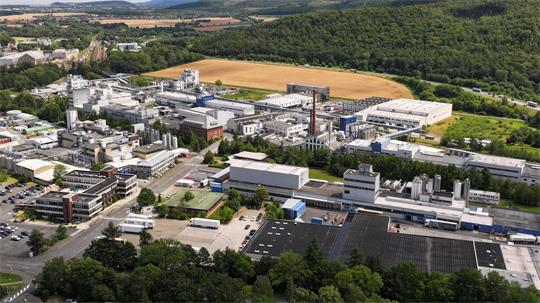 Luftaufnahme eines großen Industriekomplexes mit mehreren Fabrikgebäuden, Schornsteinen und umgebenden Feldern sowie Waldflächen