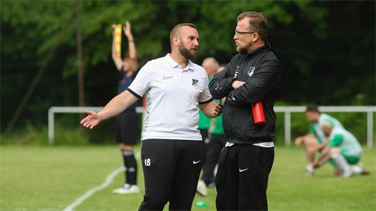 Zwei Männer in Sportkleidung unterhalten sich am Spielfeldrand eines Fußballplatzes, im Hintergrund ein Schiedsrichter mit Fahne und ein Spieler in grüner Kleidung.