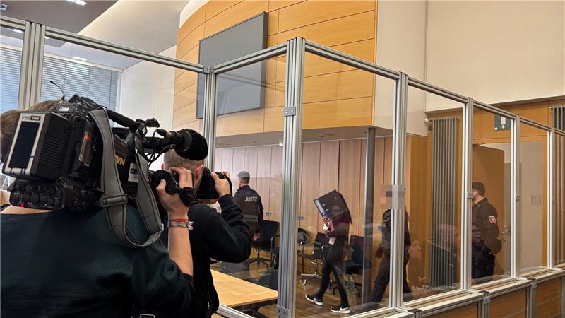 Einblick in einen Gerichtssaal: Eine Frau mit Aktenordner vor dem Gesicht betritt den Raum. Fernsehkameras sind auf sie gerichtet. Eine Glaswand trennt die Zuschauer und Journalisten von der Verhandlung.