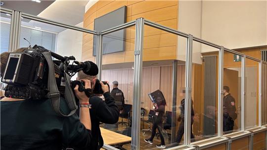 Einblick in einen Gerichtssaal: Eine Frau mit Aktenordner vor dem Gesicht betritt den Raum. Fernsehkameras sind auf sie gerichtet. Eine Glaswand trennt die Zuschauer und Journalisten von der Verhandlung.