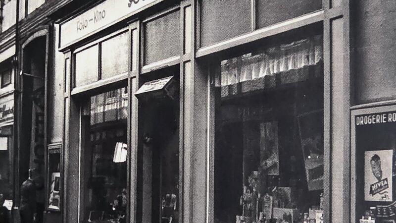 Fassade eines Drogerieladens mit Schaufenstern und Schriftzug »Rochus« in einer engen Straße mit Fachwerkhäusern.