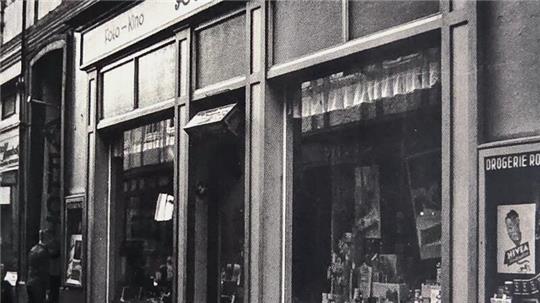 Fassade eines Drogerieladens mit Schaufenstern und Schriftzug »Rochus« in einer engen Straße mit Fachwerkhäusern.