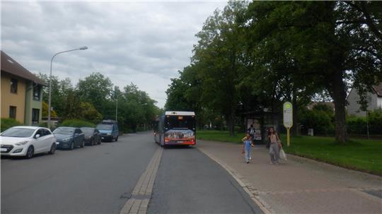 Straße mit parkenden Autos, einem Bus in der Mitte und zwei Fußgängerinnen an einer Bushaltestelle unter bewölktem Himmel.
