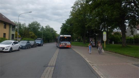 Bus fährt auf einer Straße mit mehreren parkenden Autos, zwei Personen gehen an einer Bushaltestelle entlang