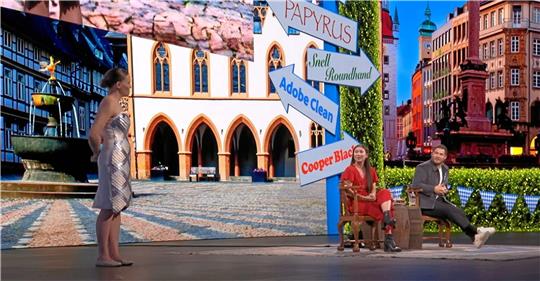 Eine Entwicklerin (links) präsentiert auf der weltweit größten Kreativkonferenz „Adobe Max“ in Los Angeles ein Kleid, das auf Knopfdruck Farbe und Muster wechselt. Im Hintergrund der Bühne ist der Goslarer Marktplatz zu erkennen. Screenshot: Youtube