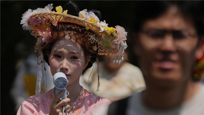 Eine Frau, die einen Hut mit Blumen und ein traditionelles Kostüm trägt, kühlt sich mit einem tragbaren elektrischen Ventilator ab, während Menschen an einem heißen Tag die Verbotene Stadt in Peking besuchen.