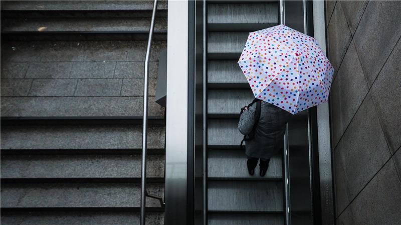 Eine Frau fährt mit ihrem Regenschirm eine Rolltreppe hinauf. Das Wetter soll in der Region Stuttgart in den nächsten Tagen wechselhaft bleiben.