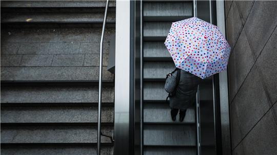 Eine Frau fährt mit ihrem Regenschirm eine Rolltreppe hinauf. Das Wetter soll in der Region Stuttgart in den nächsten Tagen wechselhaft bleiben.