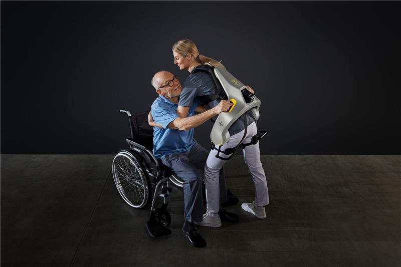 Eine Frau hebt mit Hilfe eines intelligenten Kraftanzugs der Firma German Bionic einen Mann aus einem Rollstuhl.