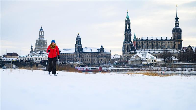 Eine Frau läuft am verschneiten Elbufer vor der historischen Altstadtkulisse in Dresden Ski